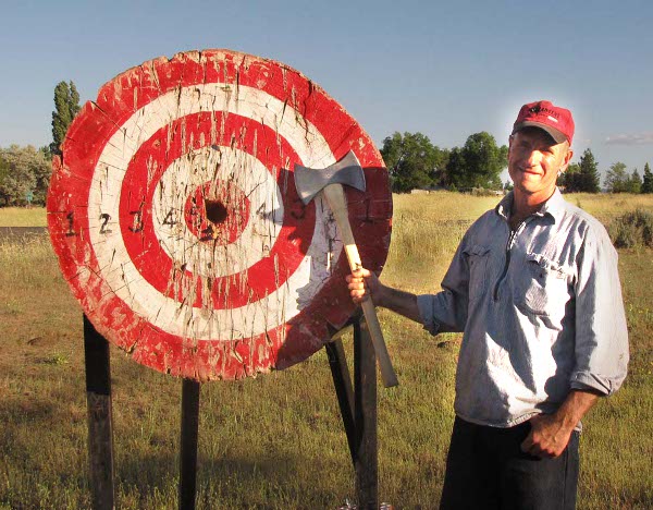 A tall thin young man stands with a throwing axe by the big wooden target.
