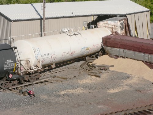 A box car half way into a steel office building with a white tank car close behind.