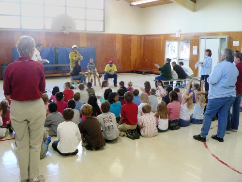 A school assembly, children sitting on floor, firemen lecturing and burn victim on chair.