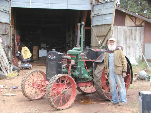 A thin old man with a white beard shows off an old stem powered tractor.