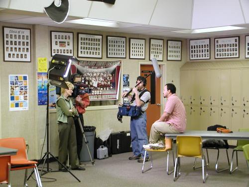 A lady interviews high school student aong with a camera man and a sound man holding a microphone boom.