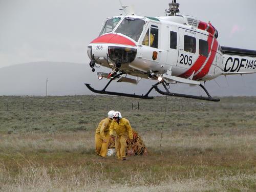 A white and Orange CDF Helicopter, aboput 3 ft off the ground, with 2 yellow clad ground crew preparing a netted pakage for pickup.