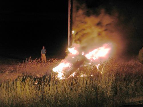 An unrecognizable object (a pickup truck) fully involved in flames and wrapped around a power pole.