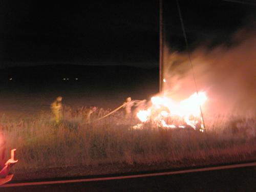 Fireman in a noght shot extinguishing a blazing vehicle around a power pole.