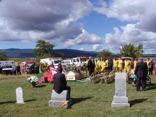 The funeral with the pine casket in the foreground and many fire trucks parked behind. Firemen in yellow uniform standing by in formation.