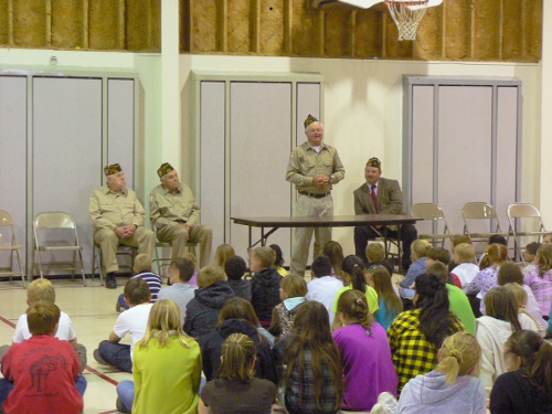Msn in VFW uniform stands in front of school children sitting on floor. He is explaining Memorial day. 3 other VFW members are seated.