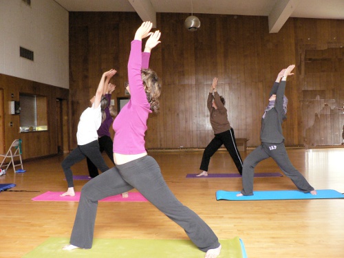 Lady with pink sweater in a sweeping yoa pose instructs her students.